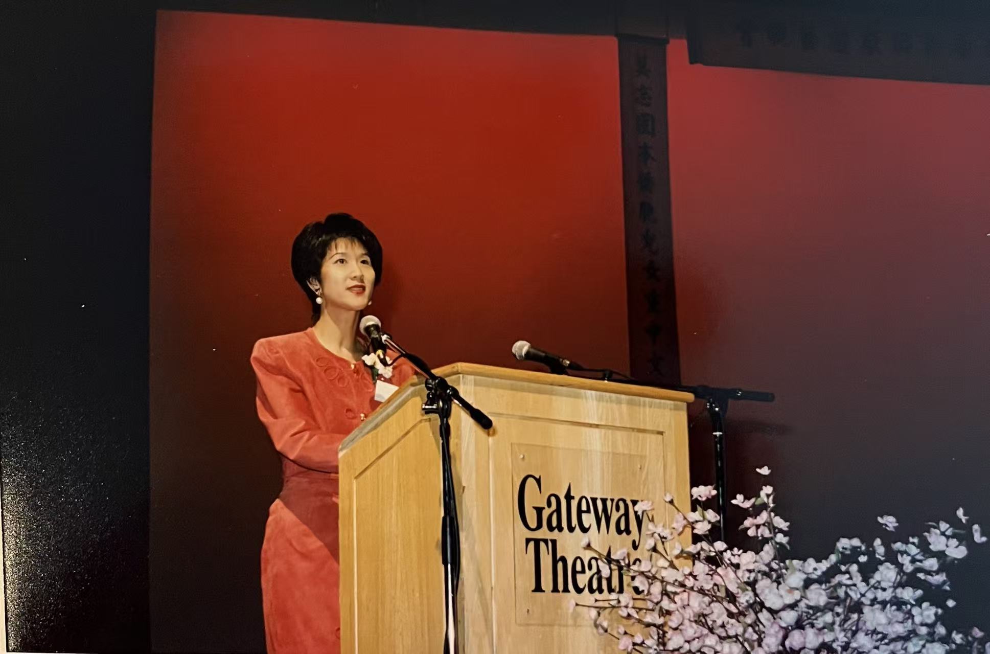 A younger M Jie-jie speaking at a podium on a stage with a red backdrop.