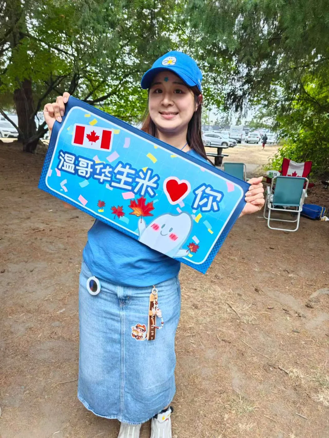 A community member holding a handmade Vancouver Shengmi sign beneath the trees.